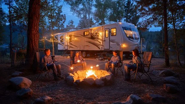 Family roasting marshmallows around campfire next to RV at dusk